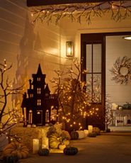 Front porch at night decorated with lit Halloween silhouettes, twig trees, hay bales, pumpkins, and candles
