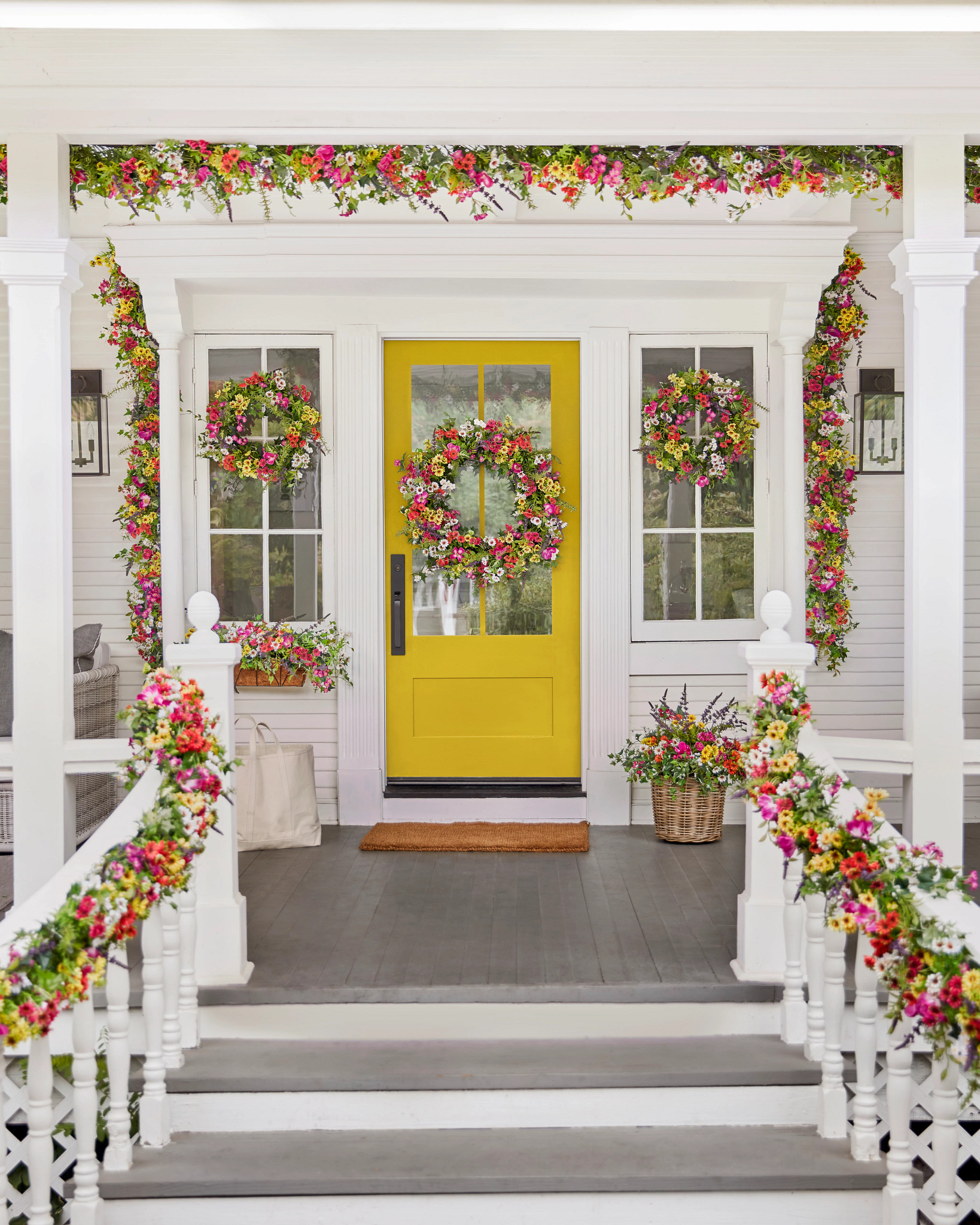 Front porch with yellow door decorated with artificial wreaths, garlands, window boxes, and potted arrangements with faux lavender, chrysanthemums, wildflowers, and mixed greenery