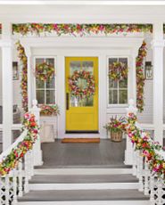 Front porch with yellow door decorated with artificial wreaths, garlands, window boxes, and potted arrangements with faux lavender, chrysanthemums, wildflowers, and mixed greenery