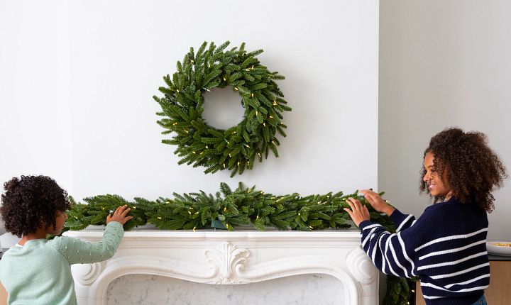 Two children decorate a white fireplace mantel with a lighted artificial wreath and garland