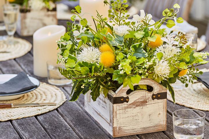Artificial floral arrangement with white and yellow flowers and green leaves in a wooden planter on a dining table with candles and place settings