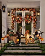 Two children in Halloween costumes stepping out of the front porch decorated with pumpkins, hay bales, fall foliage, and Jack-o’-lantern pathway lights