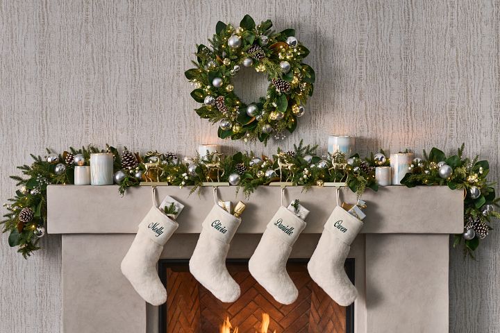 Fireplace mantel decorated with artificial greenery, candles, metallic accents, and ivory Christmas stockings