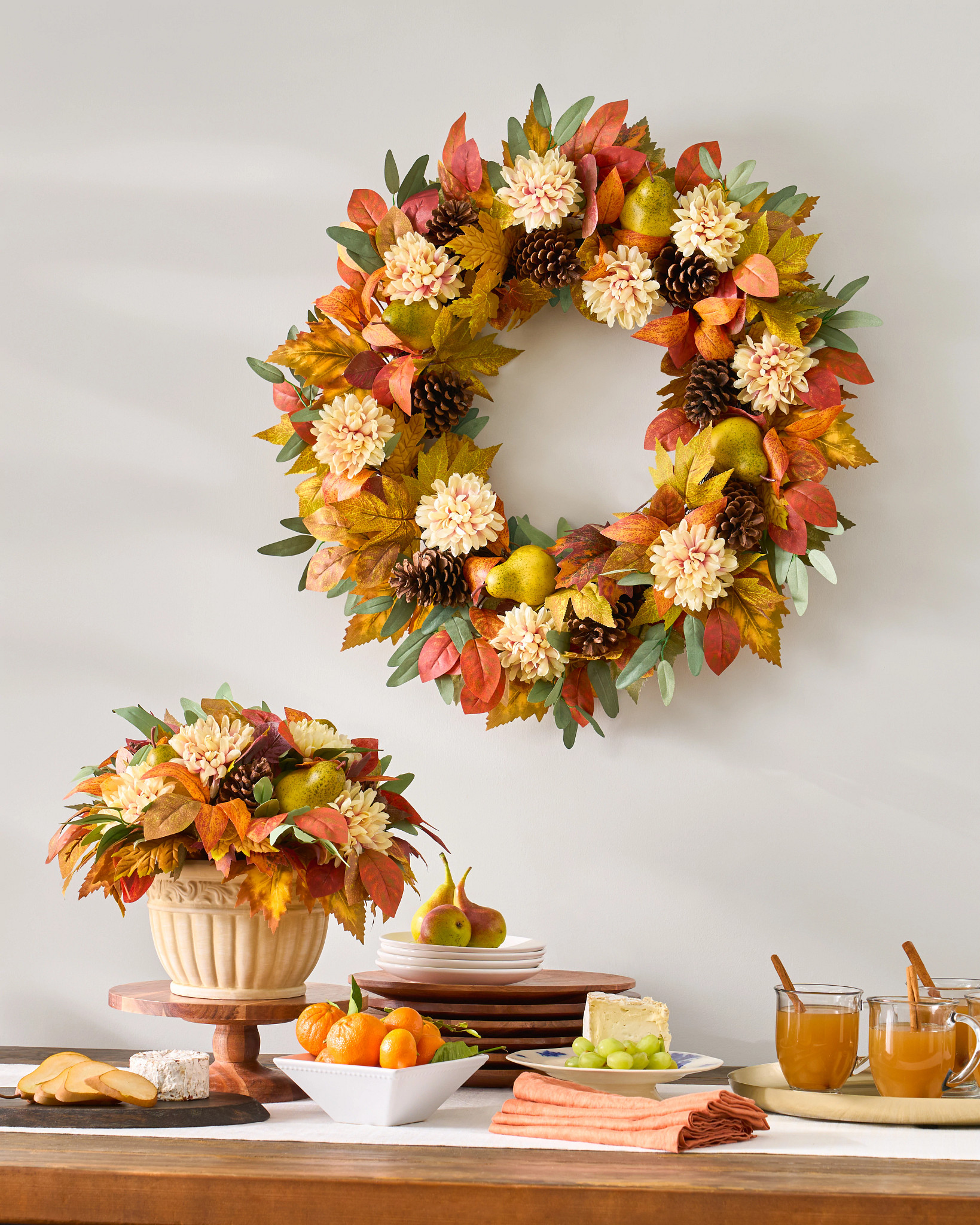 Buffet table decorated with assorted fruits and cheese, pumpkin spice drinks in clear glass mugs, and artificial wreath and floral arrangement featuring faux pears, pinecones, ivory dahlias, oak, vine maple, beech, and eucalyptus leaves