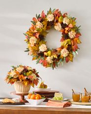 Buffet table decorated with assorted fruits and cheese, pumpkin spice drinks in clear glass mugs, and artificial wreath and floral arrangement featuring faux pears, pinecones, ivory dahlias, oak, vine maple, beech, and eucalyptus leaves