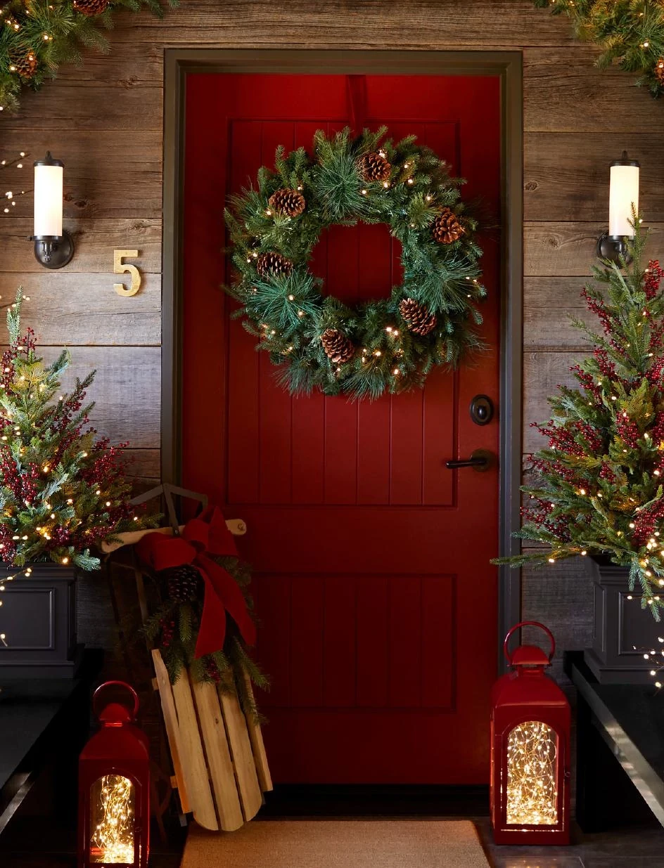 Red door with artificial Christmas wreath, sled, and lanterns