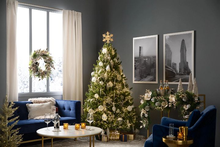 Artificial Christmas tree decorated with white and silver ornaments next to a bar cart and blue sofa in living room