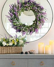 Round mirror with black frame decorated with artificial lavender wreath and dresser drawer with artificial flower arrangement and faux candles on top
