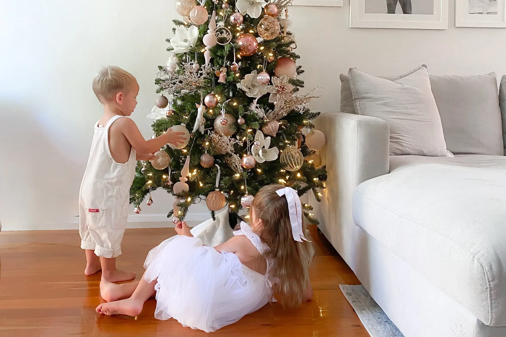 Two young children play near a beautifully decorated Christmas tree with soft lighting and ornaments, set in a cozy living room.