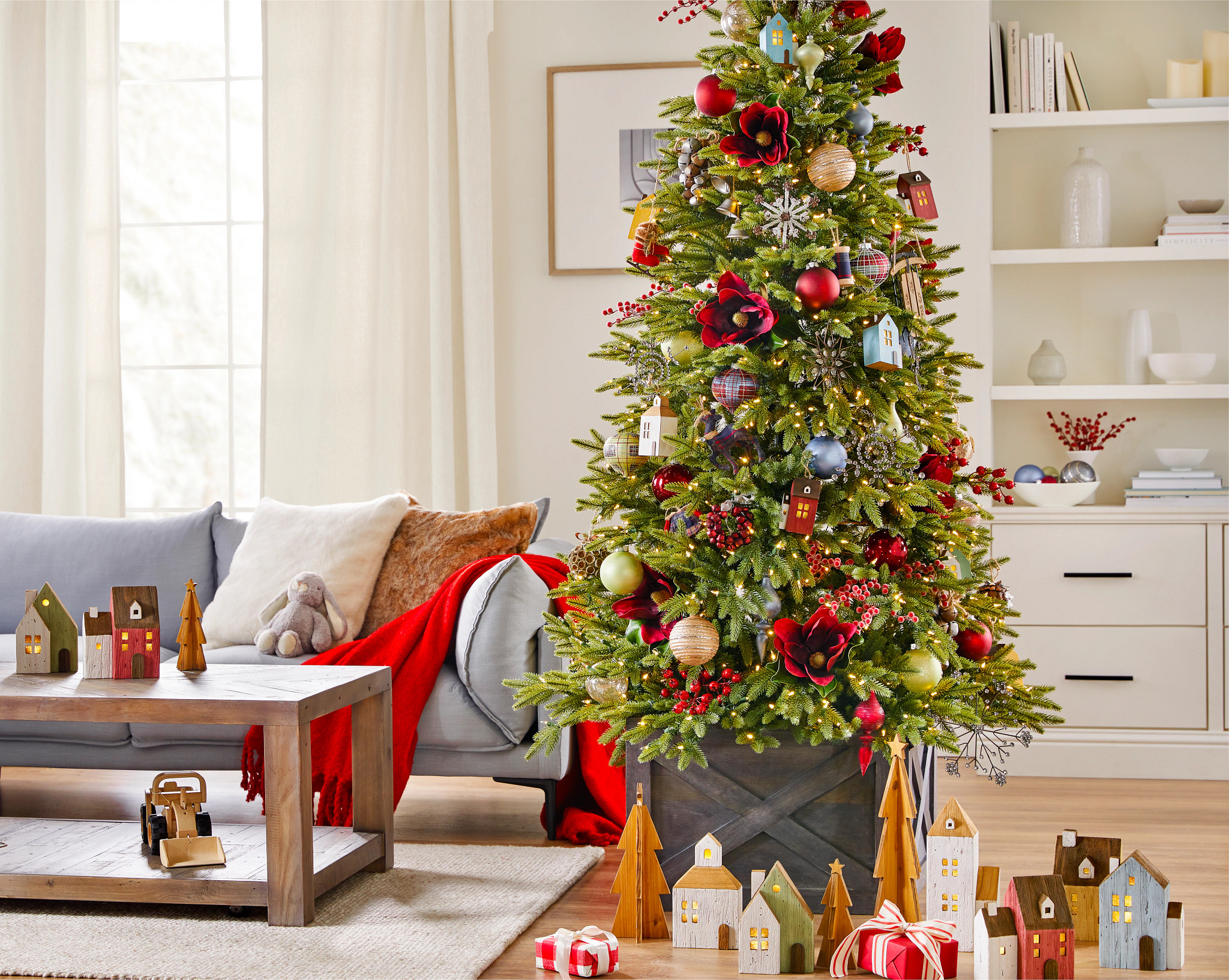 Artificial Christmas tree in living room decorated with farmhouse-themed ornaments, red magnolia stems, berry picks, and gray crate tree collar