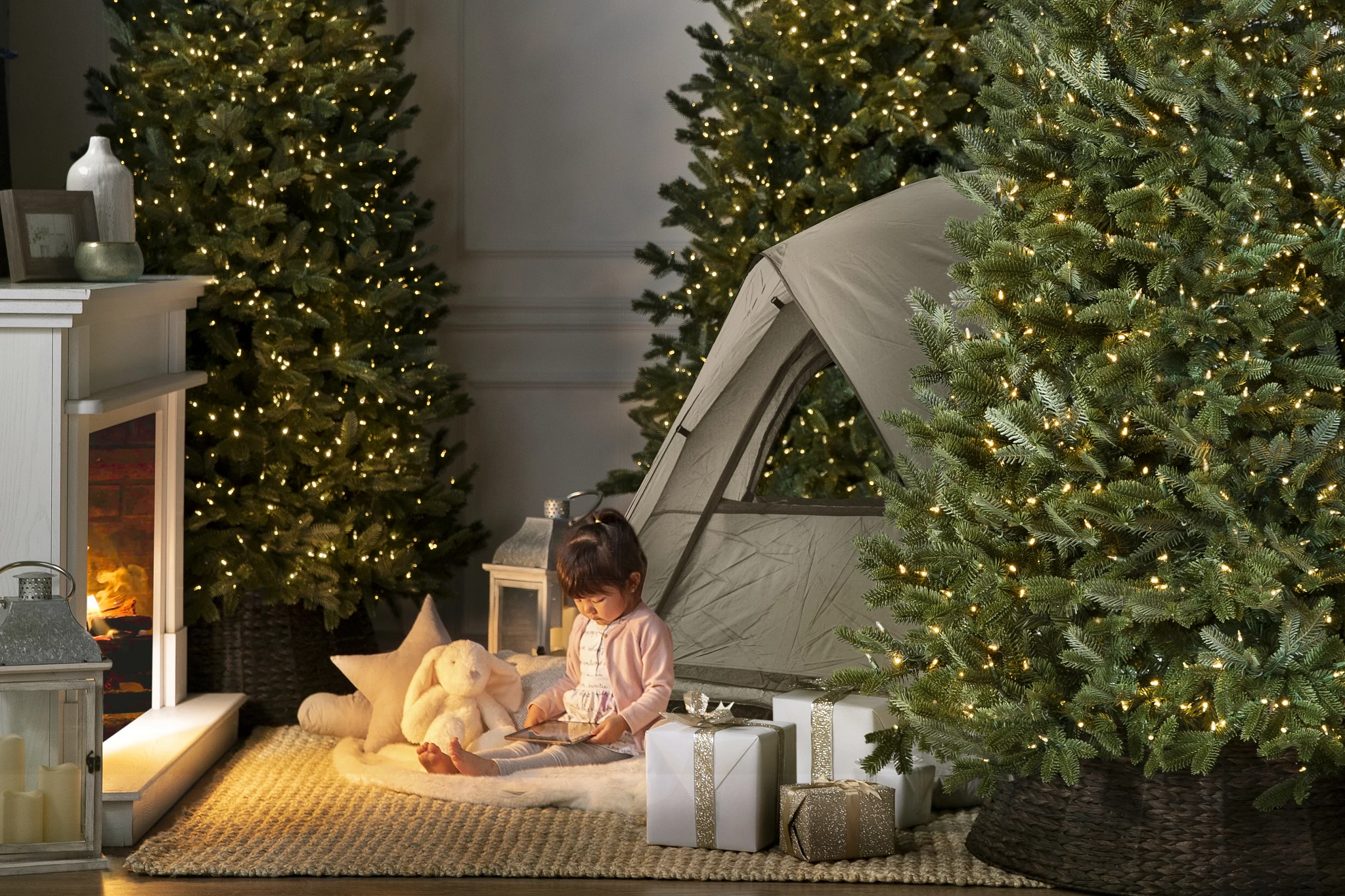 A cozy indoor scene features a tent surrounded by decorated Christmas trees, with a child and soft toys sitting on a rug near a fireplace.