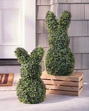 Two decorative bunny figures made of lush green artificial foliage, placed next to a wooden crate, in a garden setting.