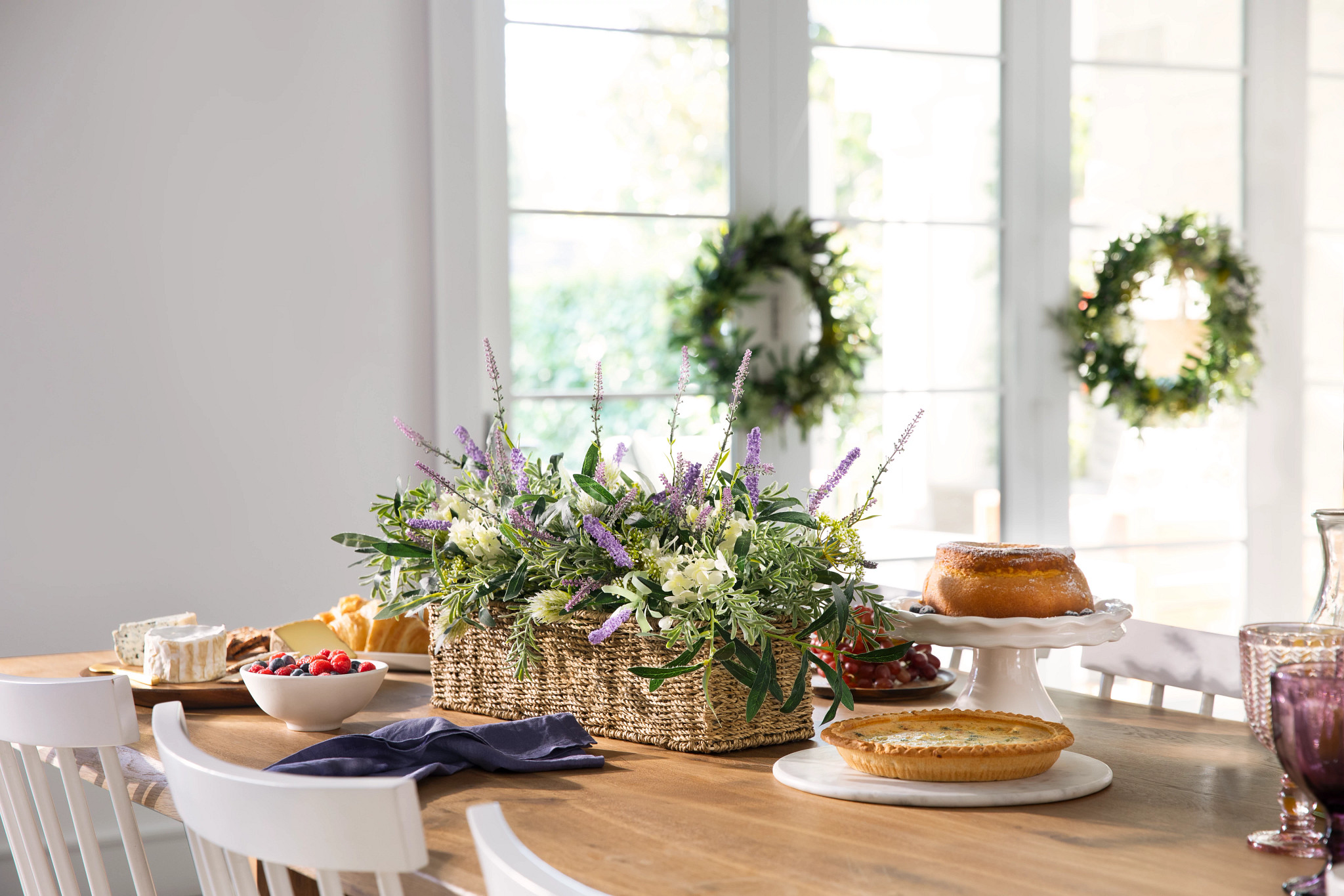 Brown dining table with an artificial flower arrangement in a rectangular basket, a bowl of mixed berries, and a cake on a white stand
