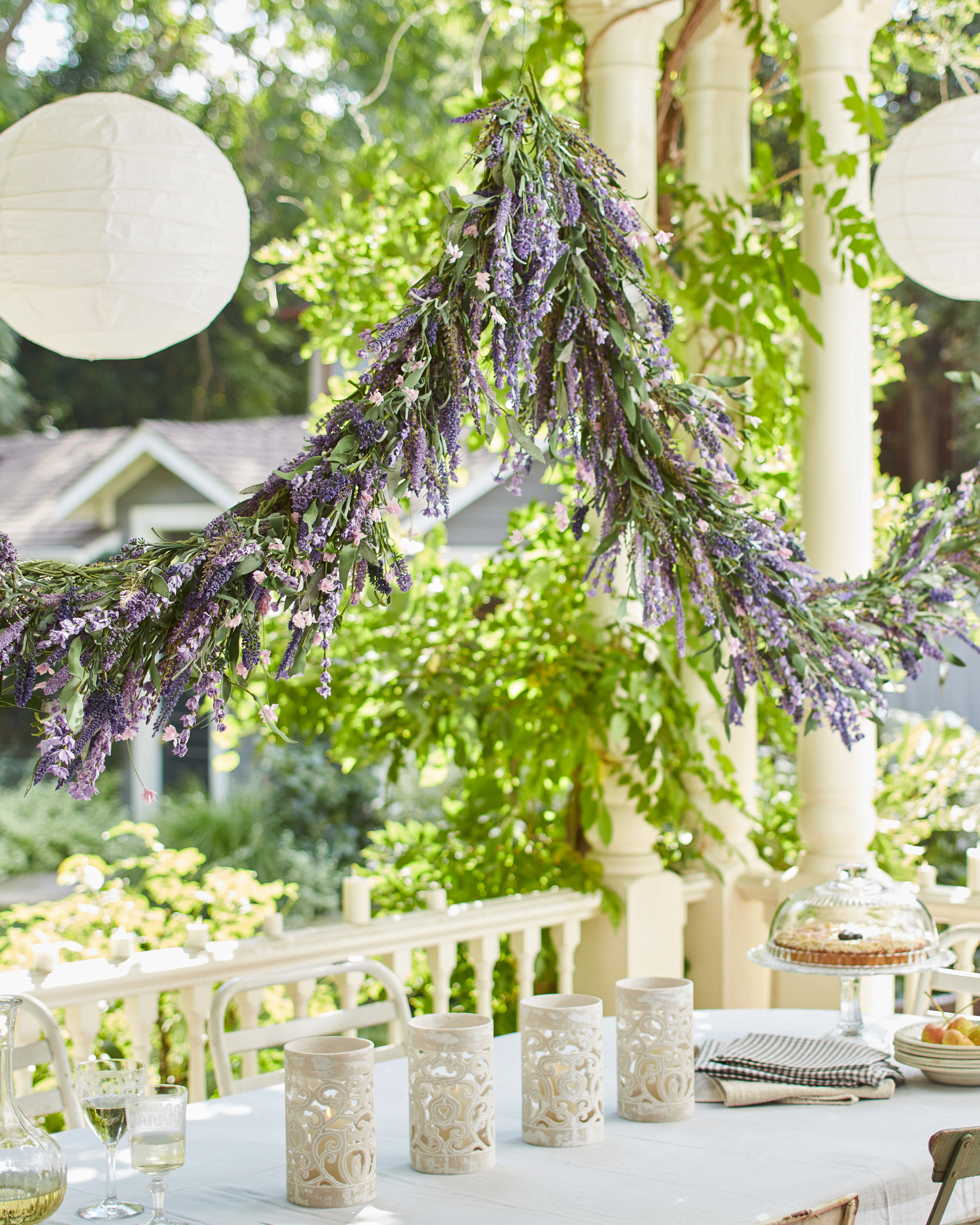 Outdoor dining area decorated with artificial lavender garland, white round paper lanterns, and white candle hurricanes