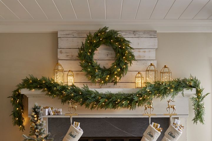 Rustic fireplace mantel décor with illuminated garland and wreath, lanterns, and white Christmas stockings