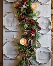 Dining table decorated with white tablecloth, place settings, faux candles, and artificial garland centerpiece with pincones, leaves, and red berries