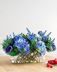 Artificial flower arrangement with faux cornflowers, anemones, hydrangeas, hypericum berries, muscari, and dusty miller leaves set in a glass vase with acrylic water illusion
