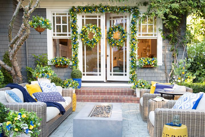 Outdoor seating area and fire pit with patio entryway decorated with blue and yellow artificial floral wreaths, garlands, hanging baskets, window boxes, and potted arrangements