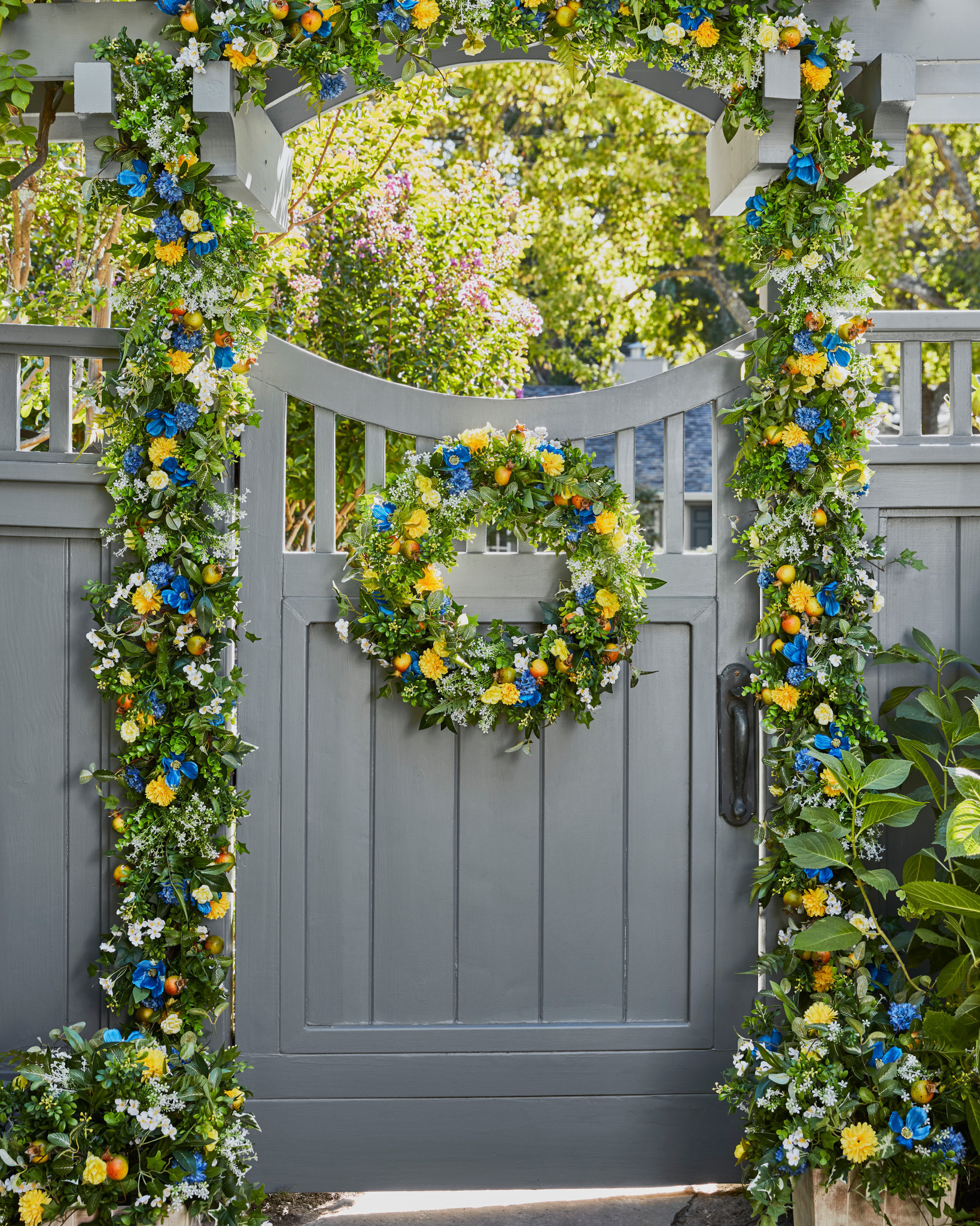 Gray wooden gate and fence decorated with artificial wreath and garland featuring faux gerberas, cornflowers, wildflowers, ivy, pomegranates, and assorted leaves