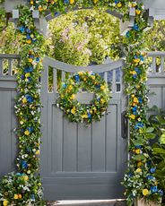 Gray wooden gate and fence decorated with artificial wreath and garland featuring faux gerberas, cornflowers, wildflowers, ivy, pomegranates, and assorted leaves