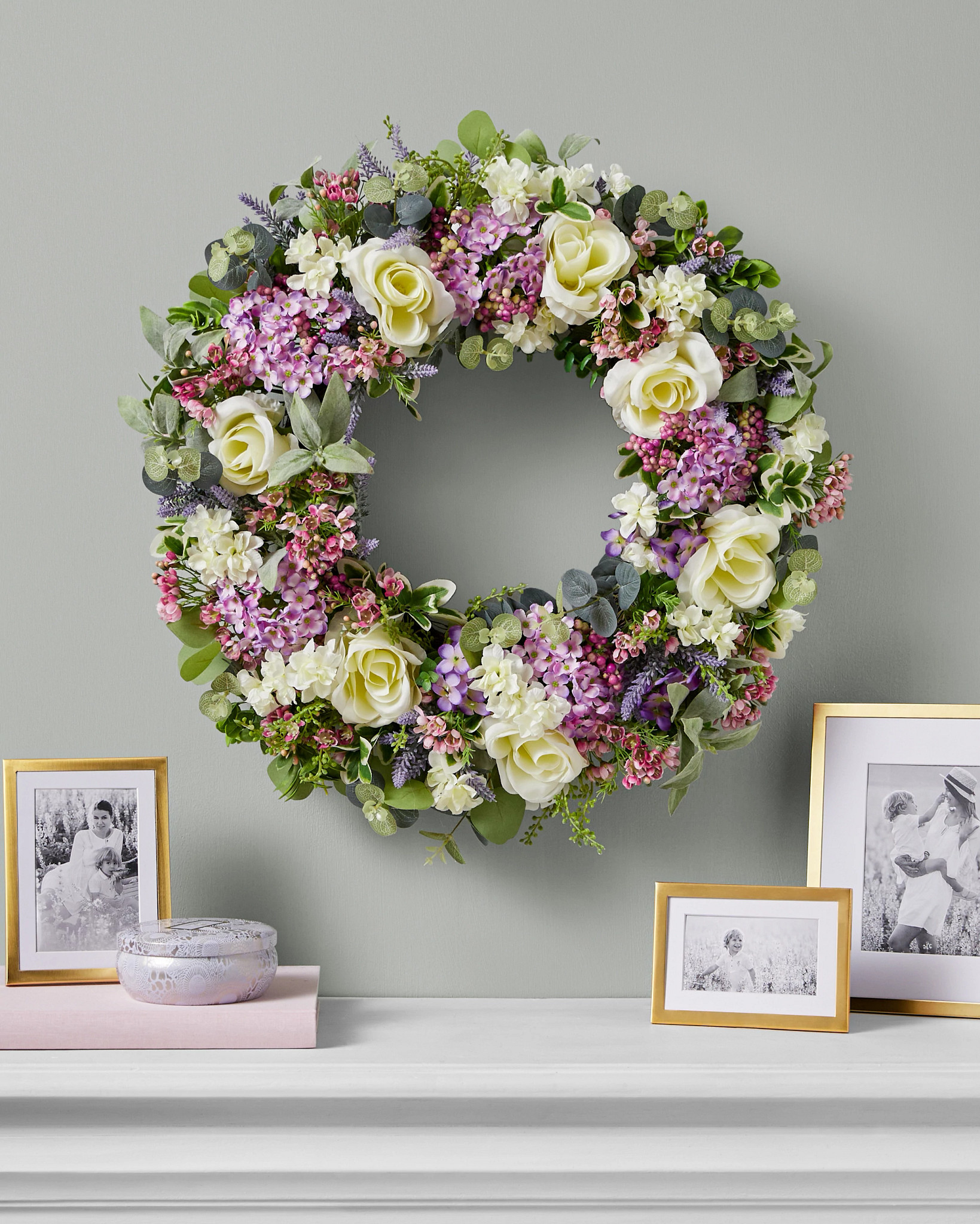 Artificial flower wreath with lilacs, roses, lavender, and hydrangeas on wall atop a white fireplace mantel