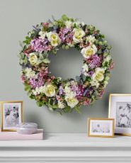 Artificial flower wreath with lilacs, roses, lavender, and hydrangeas on wall atop a white fireplace mantel