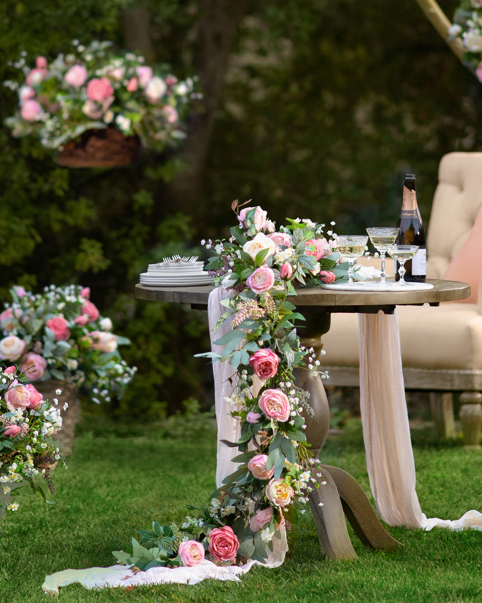 Outdoor wedding table decorated with artificial rose garland, champagne bottles and glasses, and serveware