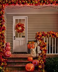 Front porch at night decorated with artificial fall greenery, lit stacked pumpkins, and a friendly ghost sculpture holding a Jack-o’-lantern