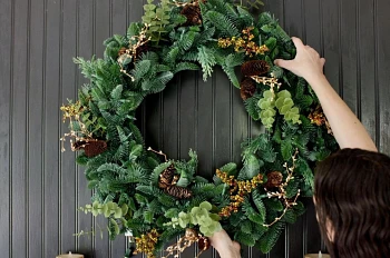 Woman hanging artificial Christmas wreath on black wall
