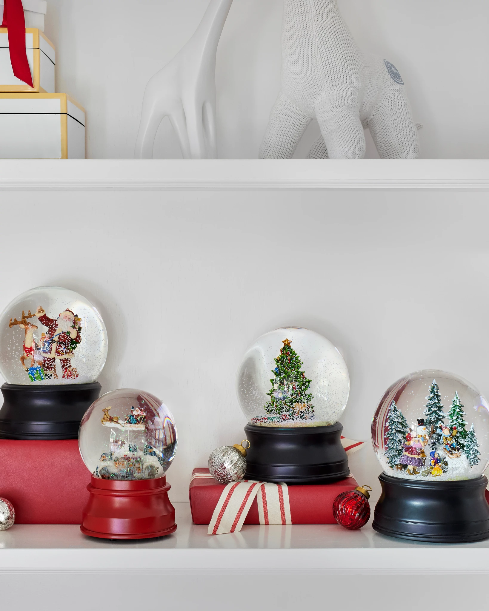 Four festive snow globes featuring Christmas scenes, displayed on stacked red gift boxes, beside ornamental decorations.