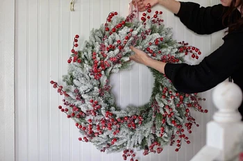 Woman hanging frosted artificial Christmas wreath with ribbon from a hook on the wall