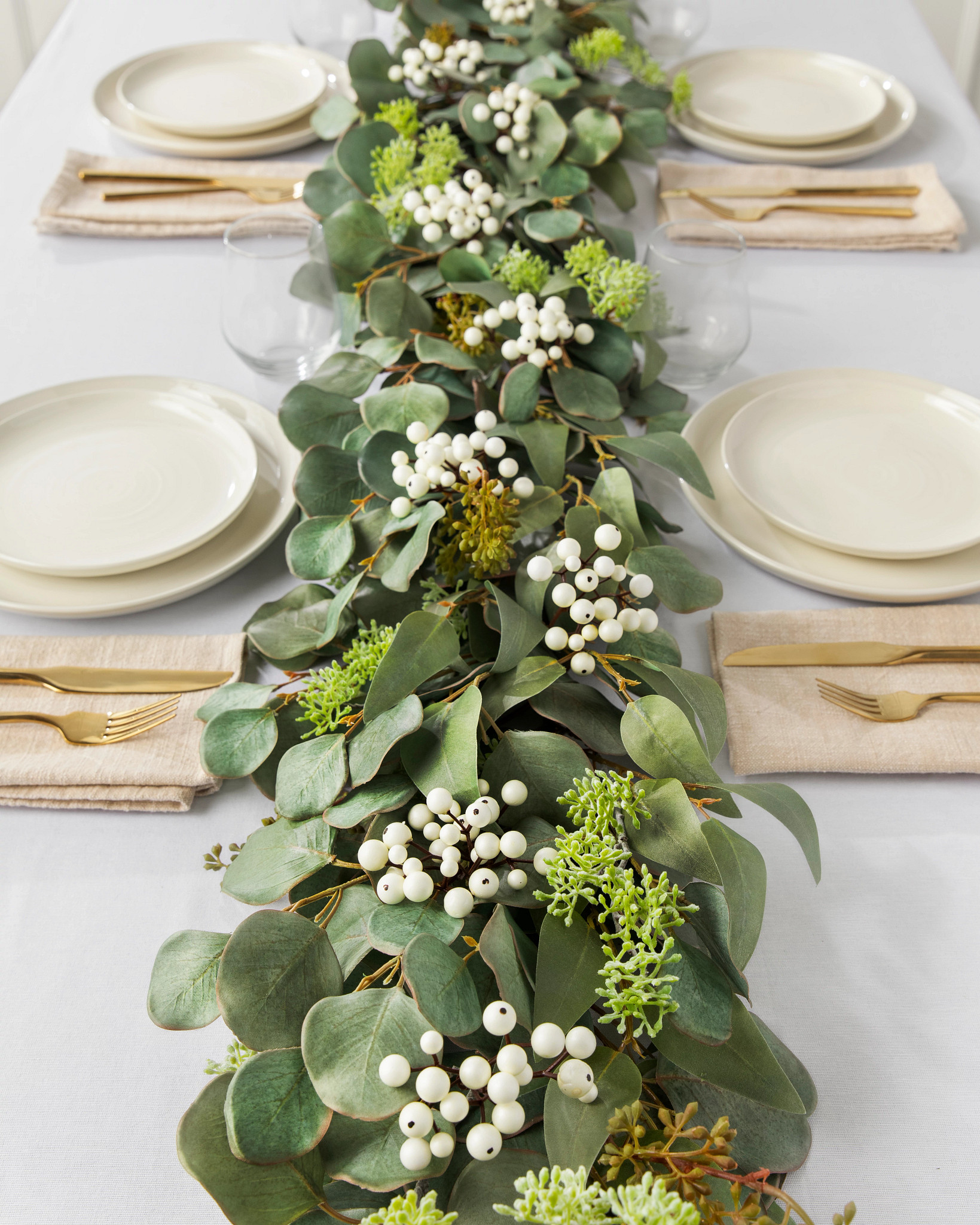 Dining table with white tablecloth, place settings, and artificial garland centerpiece featuring seeded, silver dollar, willow eucalyptus leaves, and ivory berries