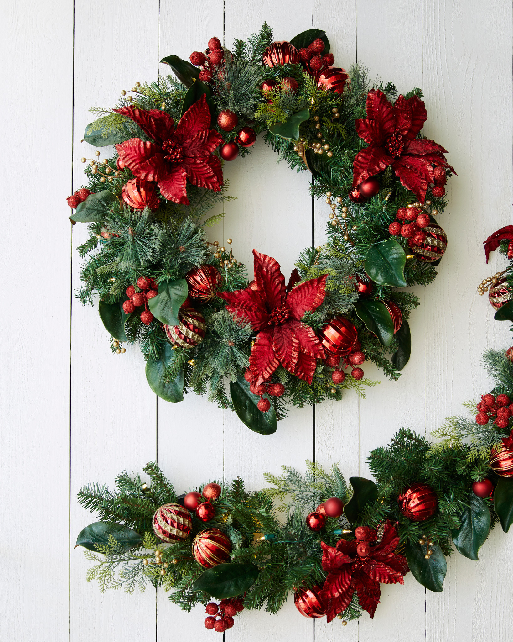 Artificial Christmas wreath and garland decorated with poinsettias, berries, and ornaments on white bakcground