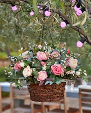 Artificial flower arrangement with faux cottage roses, eucalyptus leaves, dock stems, and ivy leaves set in a rattan basket hanging from a tree branch decorated with pink globe lights