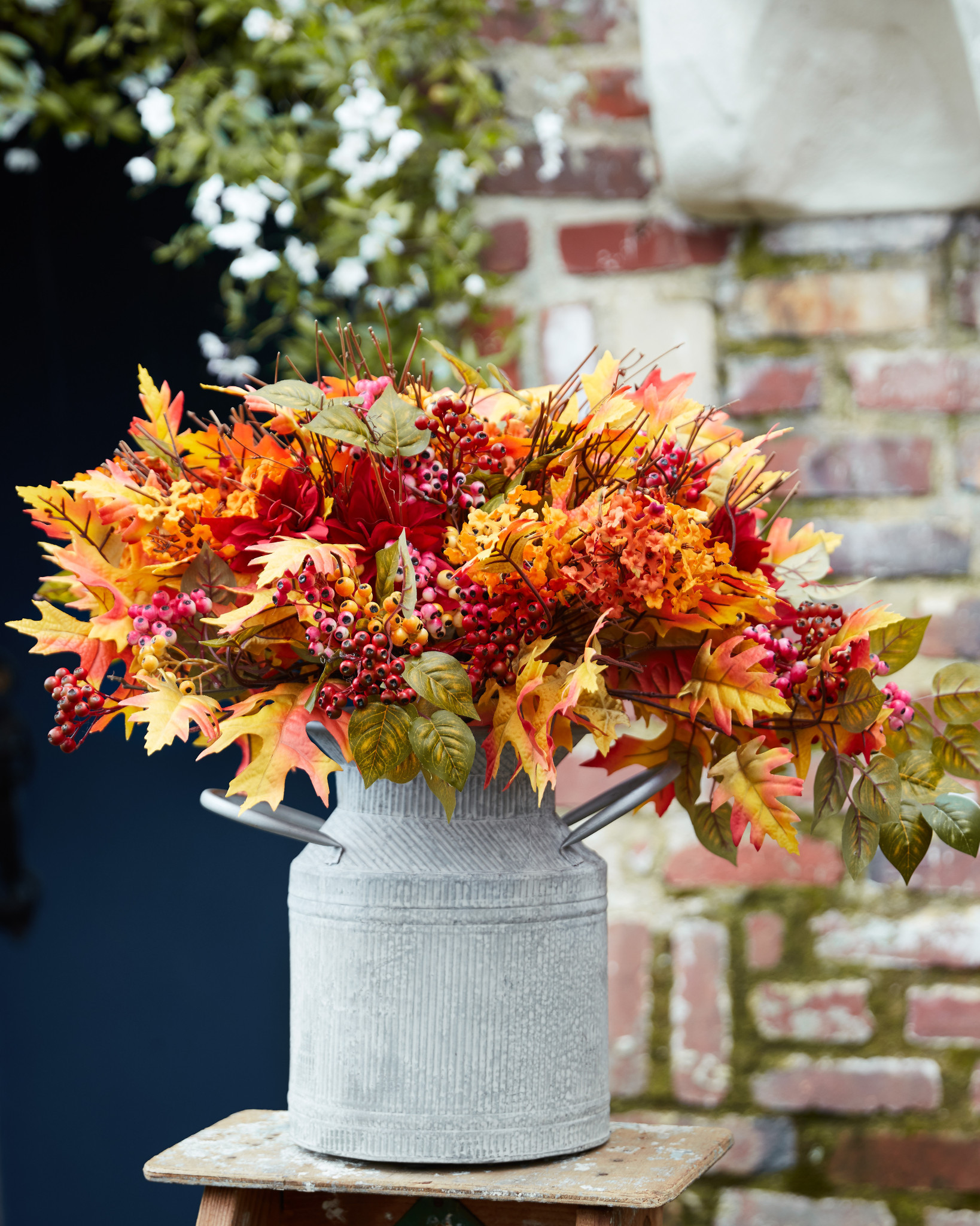 Artificial arrangement with faux golden maple leaves, magenta dahlias, and orange wildflowers set in a rustic milk can-style vessel