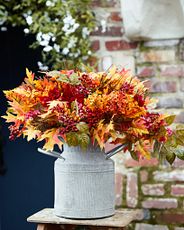 Artificial arrangement with faux golden maple leaves, magenta dahlias, and orange wildflowers set in a rustic milk can-style vessel