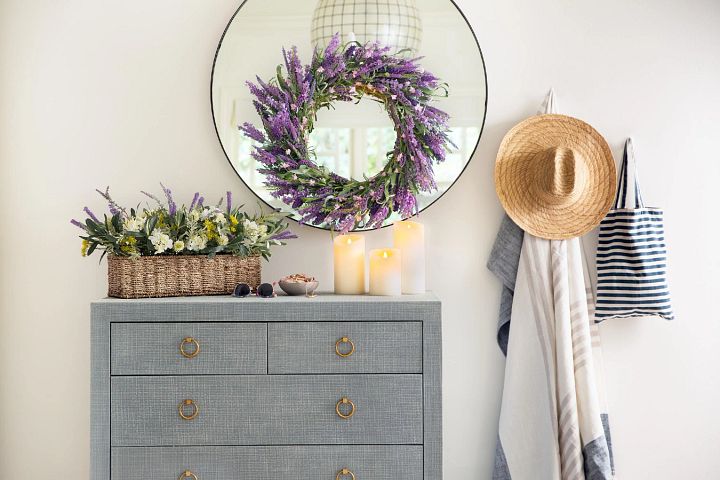 Artificial lavender wreath on round mirror and gray cabinet drawer decorated with artificial flower arrangement and candles