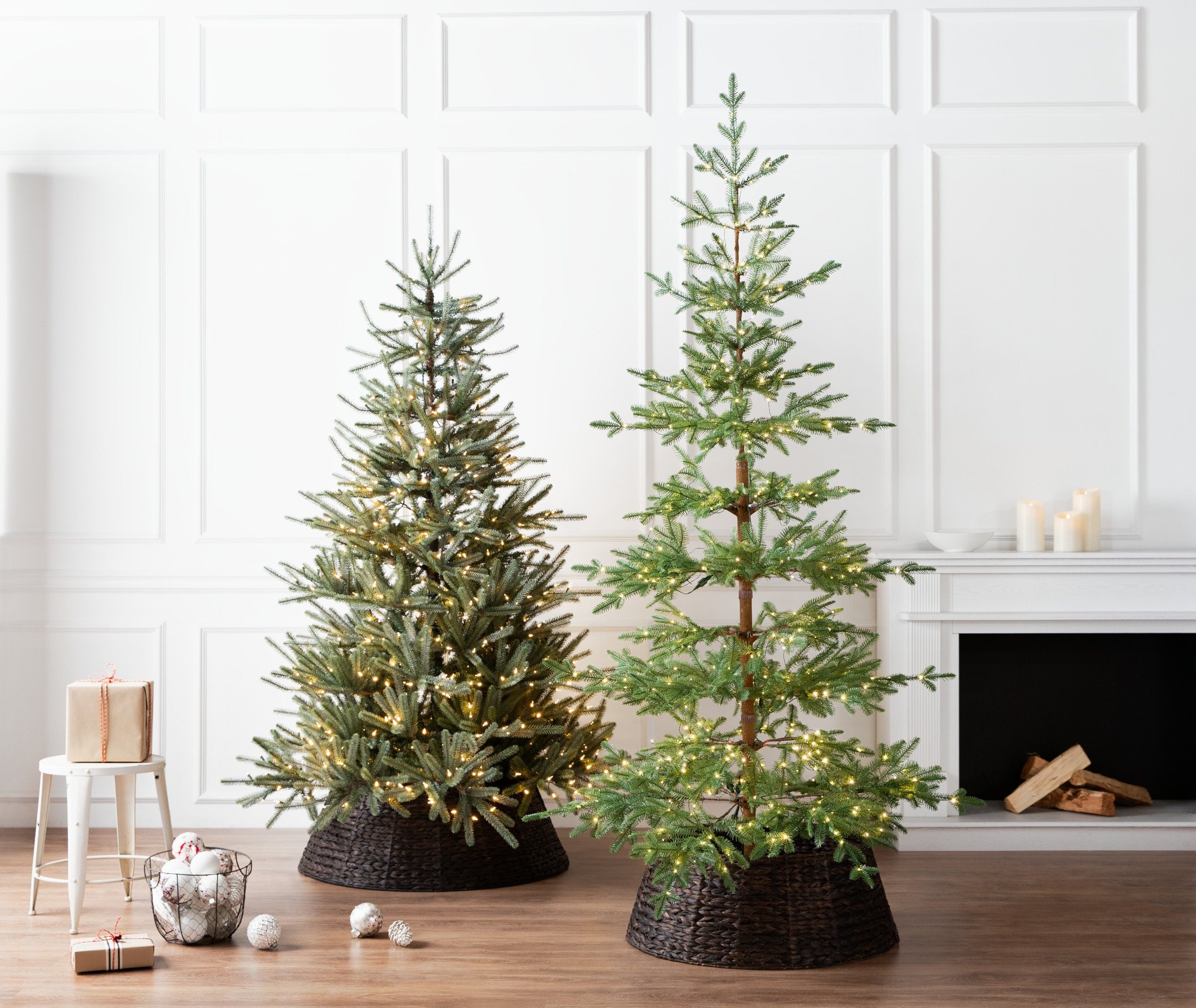 Two pre-lit artificial Christmas trees displayed in dark brown tree collars next to a fireplace in the living room