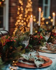 Outdoor dining table with lit candlesticks, place settings, Christmas ornaments, and artificial garland centerpiece featuring faux magnolia leaves, succulents, red berries, and natural pinecones