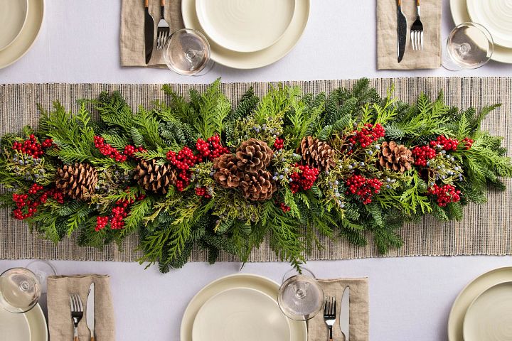 Fresh cedar garland decorated with red berries and pinecones on dining table with place settings