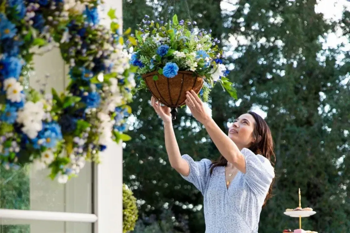 Woman hanging a blue and white artificial flower basket on a porch surrounded by lush greenery.