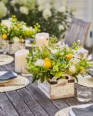 Outdoor dining table decorated with flameless candles, place settings, and artificial floral arrangements featuring angelica flowers, craspedia, and mixed greenery set in a fir wood box planter