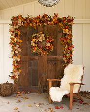 Dark wooden backdrop decorated with artificial wreath and garland featuring faux sunflowers, pumpkins, berries, and pinecones next to a wooden lounge chair with a white faux fur throw