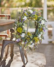 Outdoor metal dining chair decorated with artificial flower garland featuring  purple cattails, white lilacs, and thistle with mixed greenery