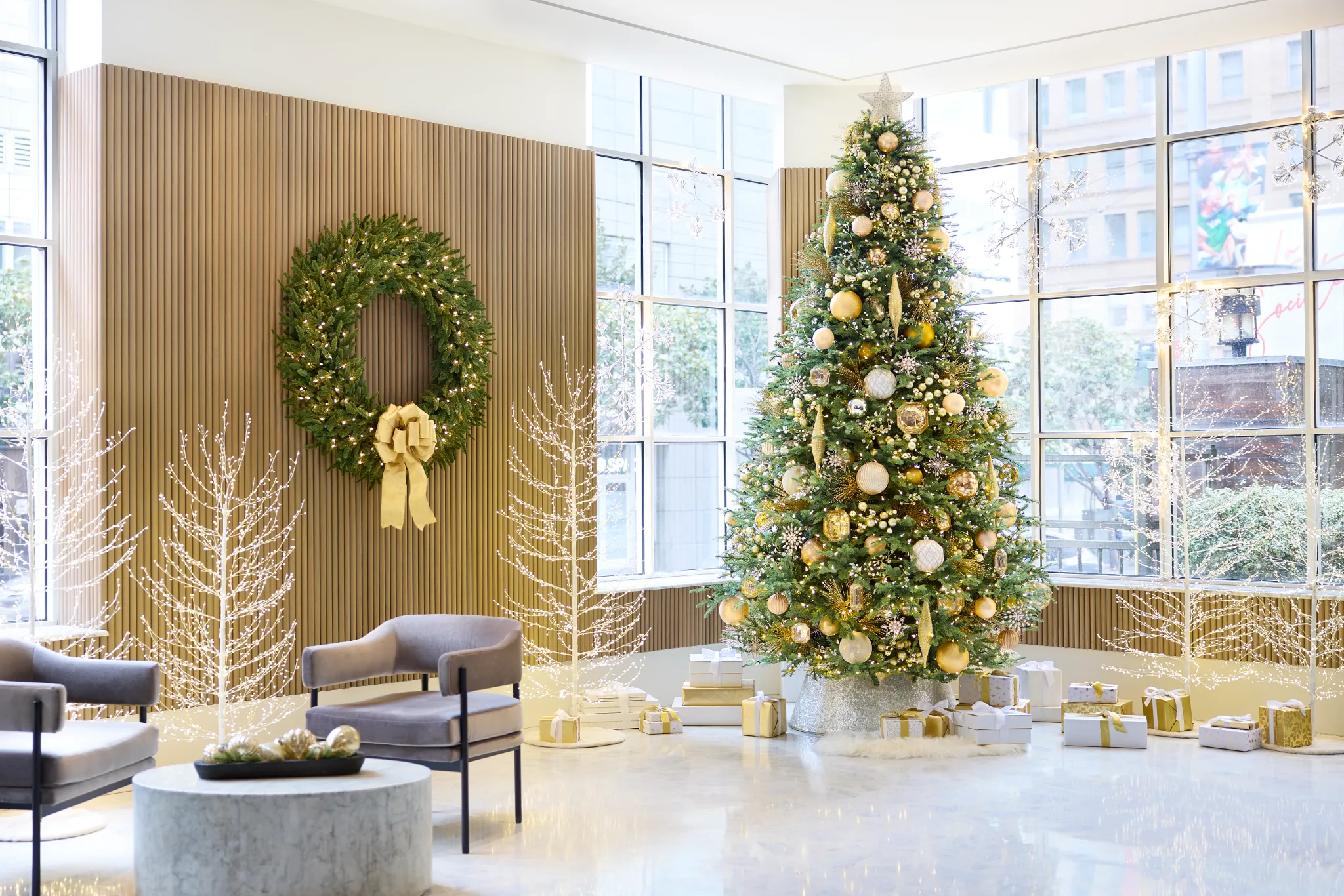 Modern lobby with large windows featuring a tall Christmas tree decorated in gold and white ornaments, a lit wreath with a gold bow, white LED twig trees, and stacked gold and white presents.