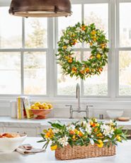Artificial wreath on window and floral arrangement on white marble countertop decorated with kumquats, lemons, orange blossoms, olives, and olive leaves