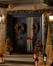 Black front door and steps decorated with artificial Christmas greenery, lanterns, and lit cone trees