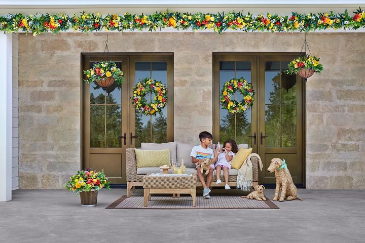 Two children sitting on wicker patio furniture decorated with colorful flowers in pots and wreaths