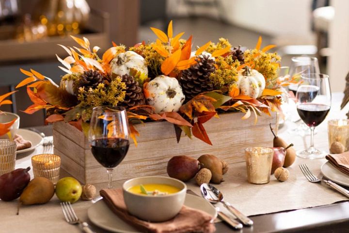 Dining table with place settings, pears, pumpkin soup in bowls, and fall centerpiece with white pumpkins, pinecones, and mixed leaves in a wooden planter