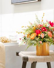 Artificial floral arrangement with cottage roses, ranunculus, peonies, billy buttons, cosmos, eucalyptus, and fern leaves in a gold ceramic pot on a stool next to bath tub